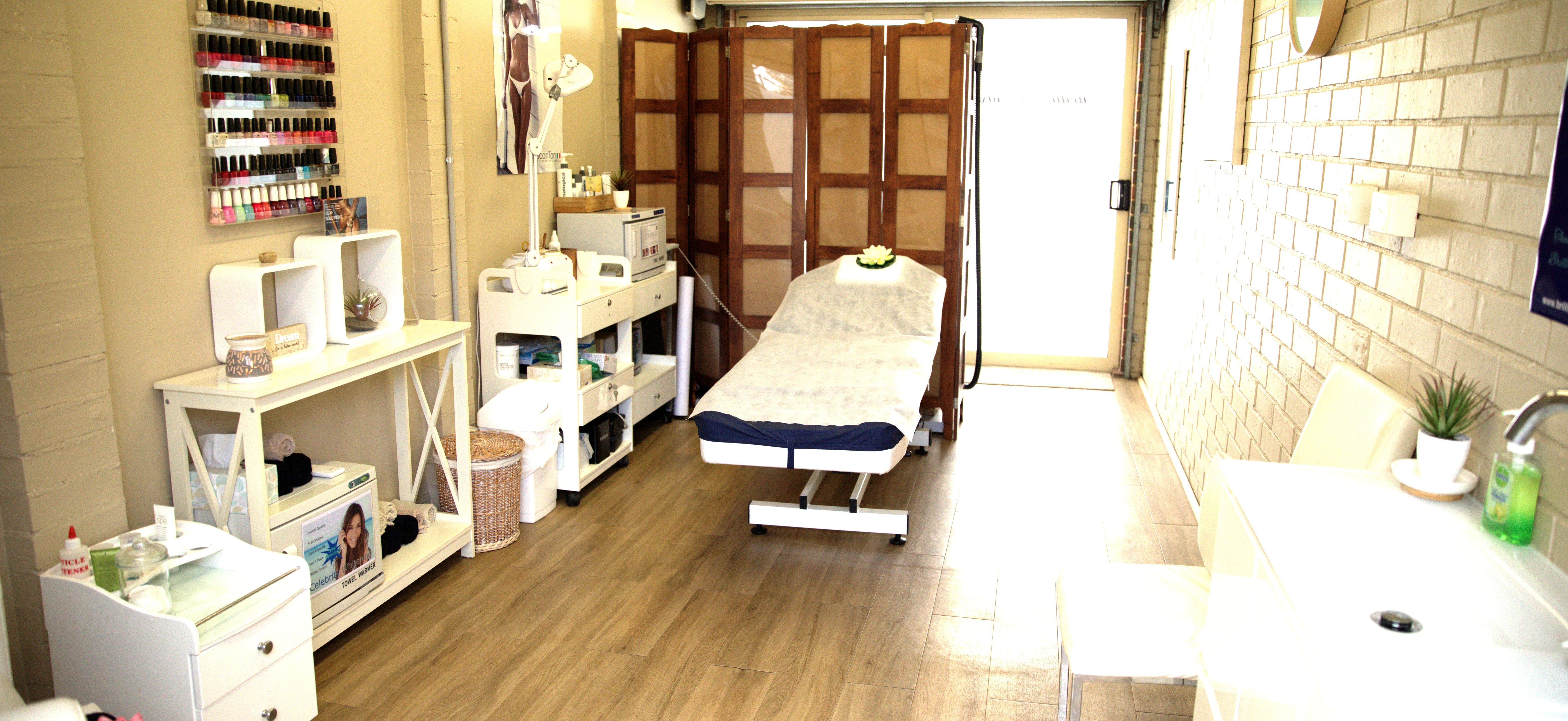 The Brilliant Beauty studio interior: treatment bed at centre, polish rack on the left wall, manicure station on the right, open doorway filled with daylight at the far end